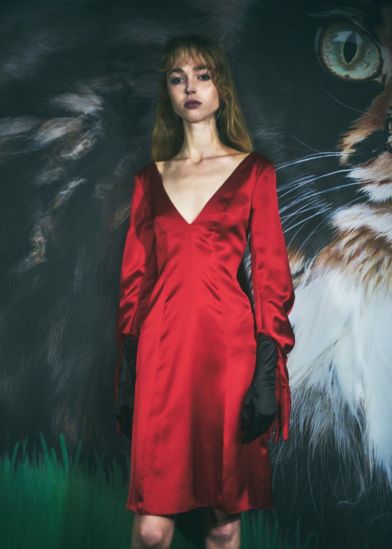 A slender, light-skinned adult female with bangs poses in a long-sleeved red dress against a backdrop featuring a giant cat face.