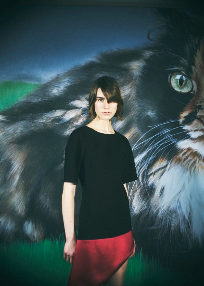 Fashion shoot of a female model with brunette hair, wearing a black top and red skirt, posing indoors in front of a cat mural.