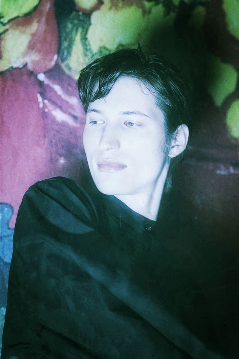 Close-up portrait of a young adult in a dark top, gazing to the side against an abstract floral background with a vintage, color-washed effect.