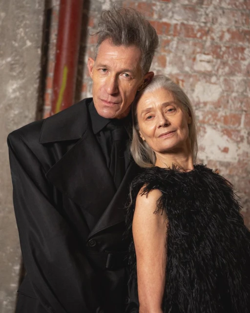 An older couple poses in front of a brick wall: the man in a black coat and the woman in a feathered top, both with grey hair.