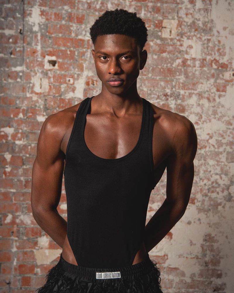 Portrait of a young Black man in a black bodysuit against a brick wall. Studio lighting, medium shot, serious expression.