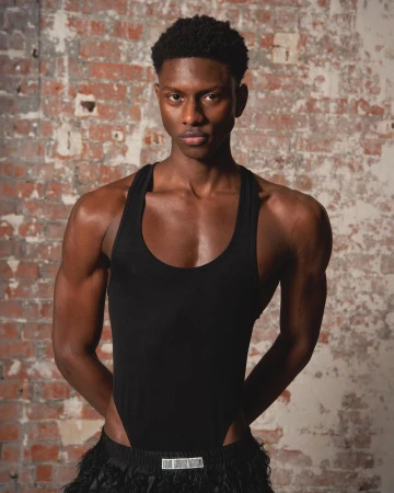 Portrait of a young Black man in a black bodysuit against a brick wall. Studio lighting, medium shot, serious expression.