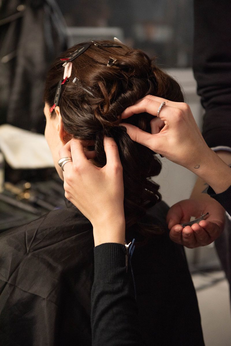 An adult female has her brunette hair styled by hands that hold hair clips in an indoor setting, indicating a hair salon or styling session.
