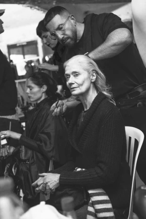 Black and white photograph of a woman sitting in a chair getting her hair styled by a hairstylist. Another woman sits in the background.