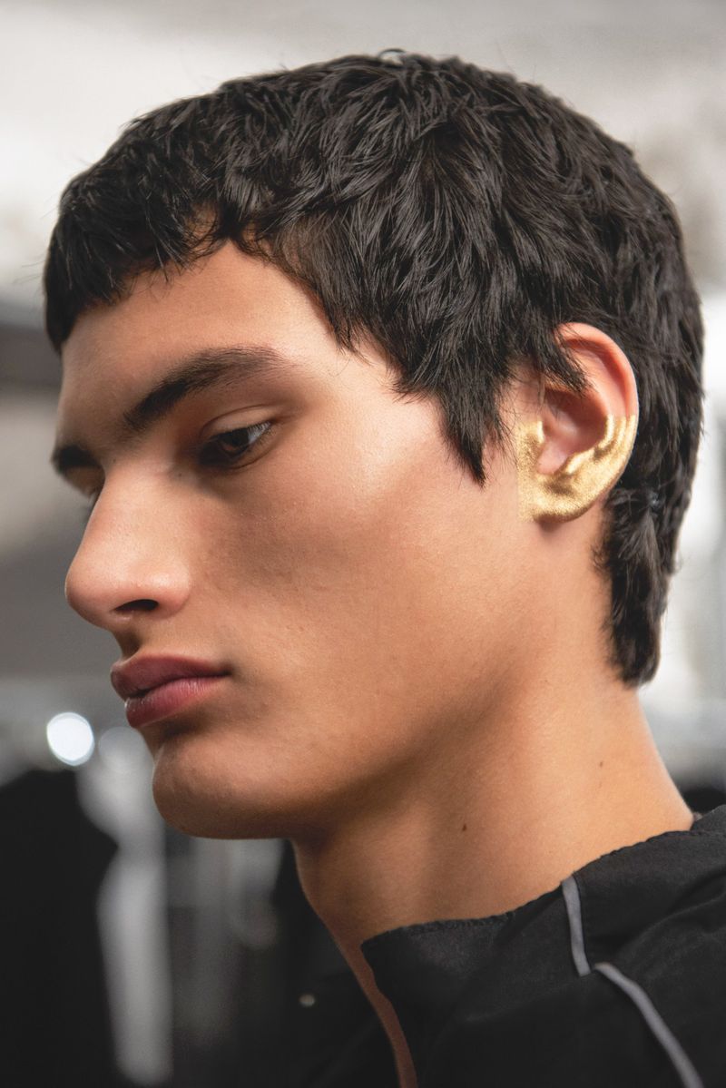 Striking close-up of a young adult male model with an expressive side profile, dark hair, and gold painted ear, in a bright setting.