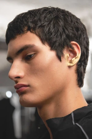 Striking close-up of a young adult male model with an expressive side profile, dark hair, and gold painted ear, in a bright setting.