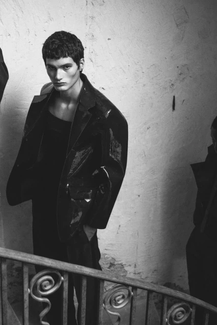 Black-and-white shot of a dark-haired male model in a black, shiny jacket and dark clothing, posing on a staircase in a dimly lit indoor location.