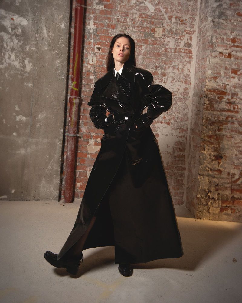 Fashion shot of a woman in a glossy black trench coat standing in an industrial urban interior setting with brick and concrete walls.