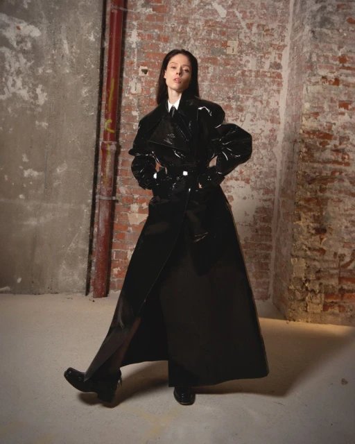 Fashion shot of a woman in a glossy black trench coat standing in an industrial urban interior setting with brick and concrete walls.