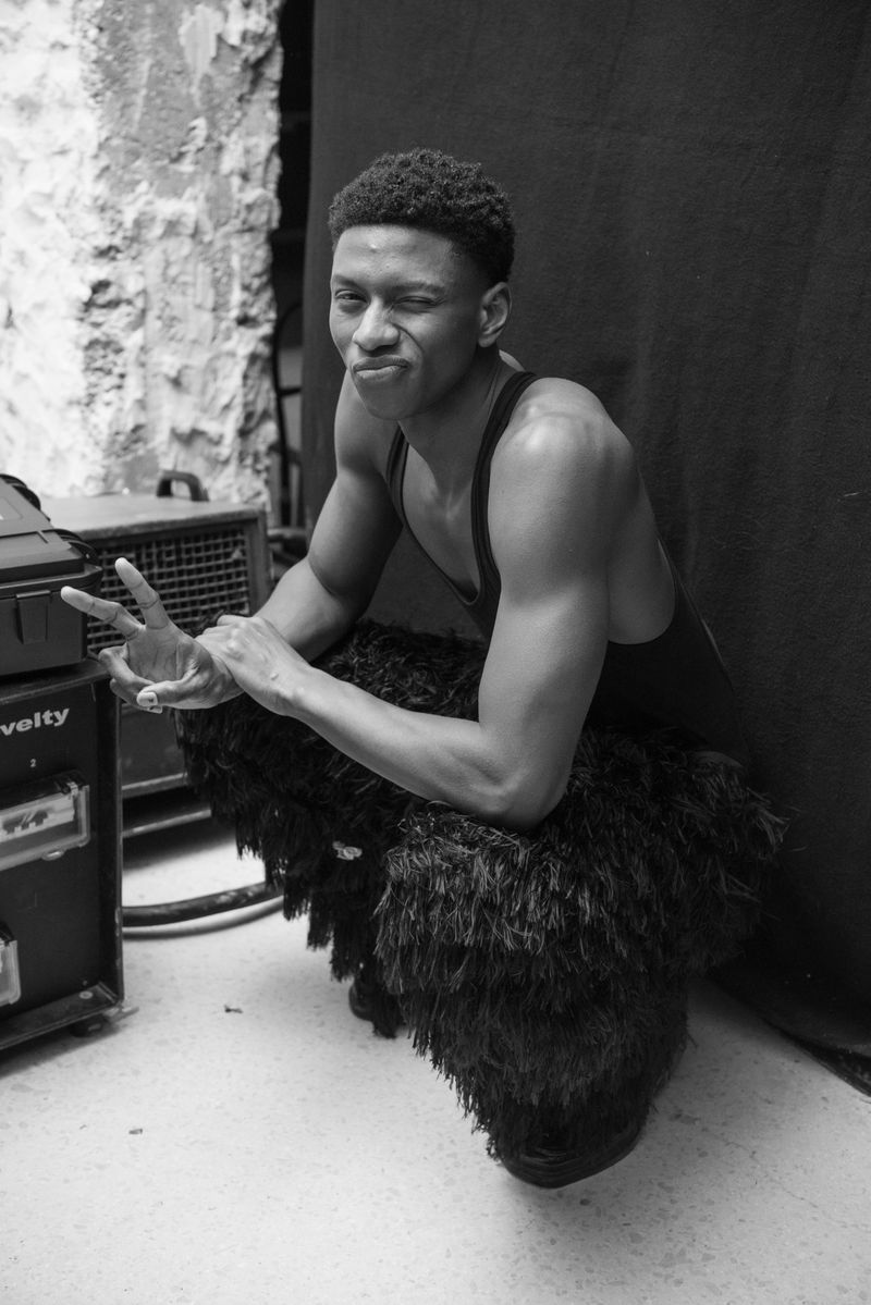 A monochrome image features a Black man in a tank top, crouching on a furry object and giving a peace sign. He is in an indoor setting.