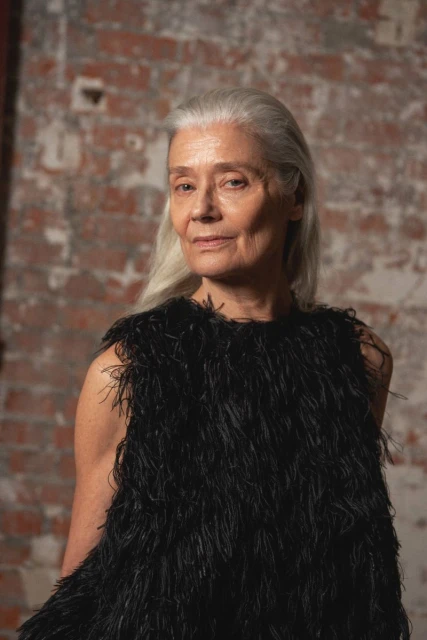 Portrait of an older woman with grey hair wearing a feathered top against a brick wall background indoors in a medium shot.