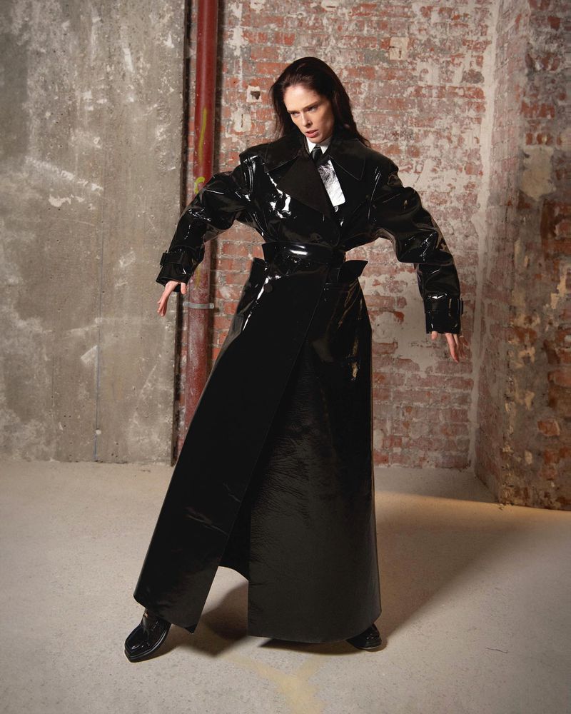 A slender adult female in a long, black patent leather coat poses in a room with concrete and brick walls, showcasing a high-fashion editorial look.