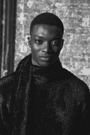 Portrait of a black man wearing a textured black sweater against an industrial brick wall background.