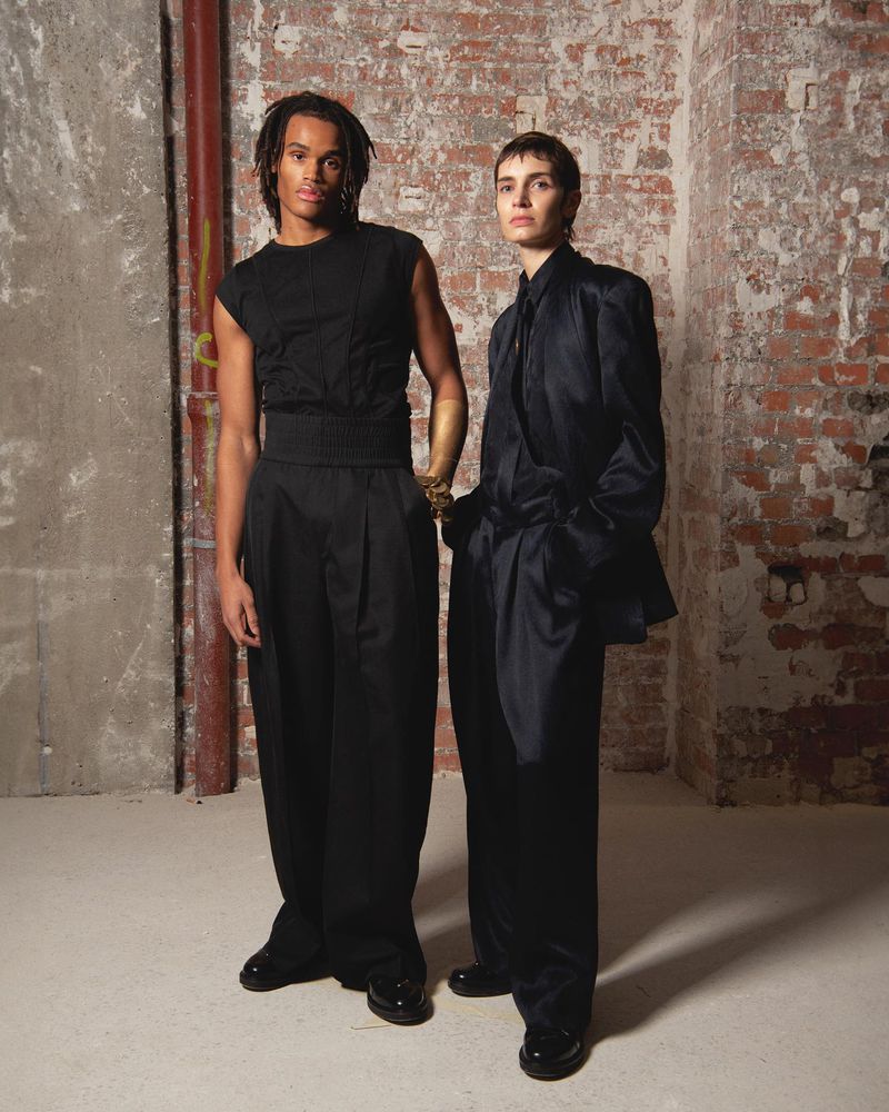 Two fashion models in black outfits pose in front of a brick wall with neutral lighting, creating a high-fashion editorial look.