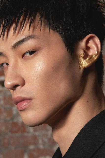 Close-up of a stylish adult male model with dark hair wearing a black shirt and an elaborate gold ear cuff against a brick wall.