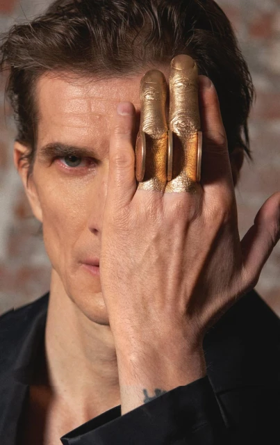 Close-up of a man with gold-painted fingers and gold rings, facing camera with partial face view and brick wall backdrop.