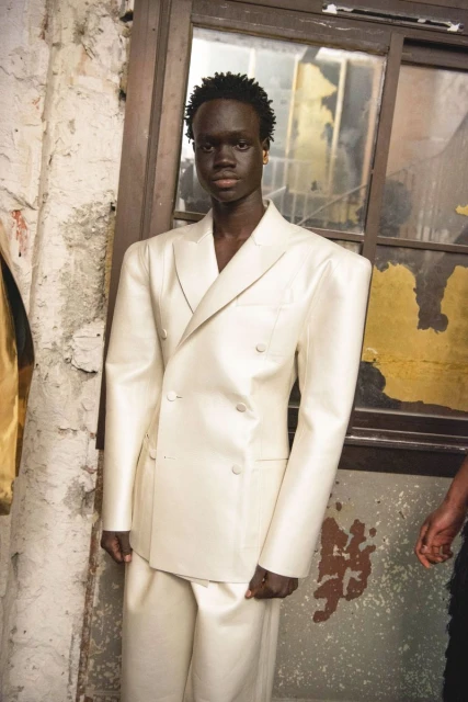 Portrait of a Black male model in an ivory double-breasted suit with padded shoulders posing in an urban indoor setting with a distressed wall.