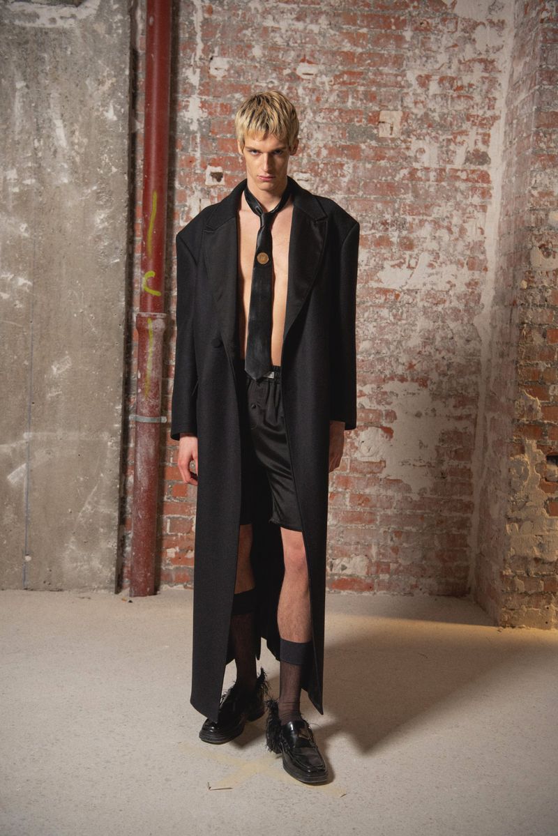 Fashionable man in an unbuttoned black coat, shorts, tie, and shiny shoes poses against a textured brick wall indoors with neutral lighting.