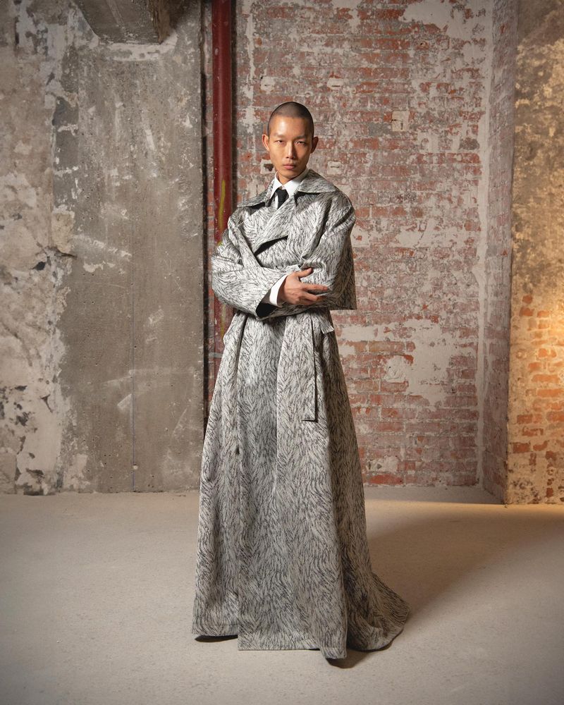 A model poses in a long gray coat in front of a distressed wall. The portrait is set indoors with neutral color tones and diffused lighting.
