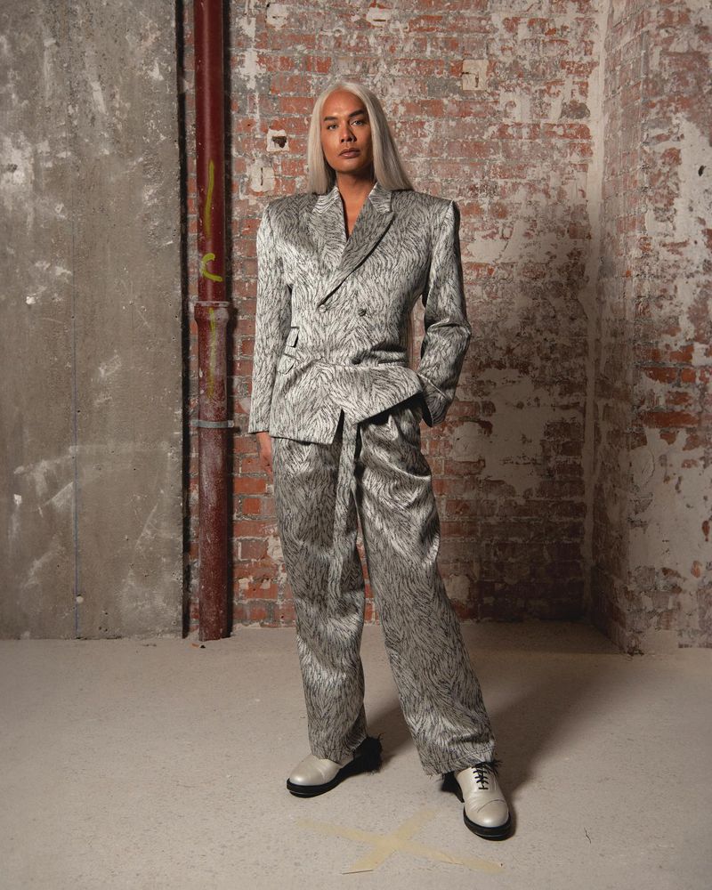 Adult in patterned suit poses in front of brick and concrete wall; silver blazer and wide leg pants with chunky lug sole shoes.
