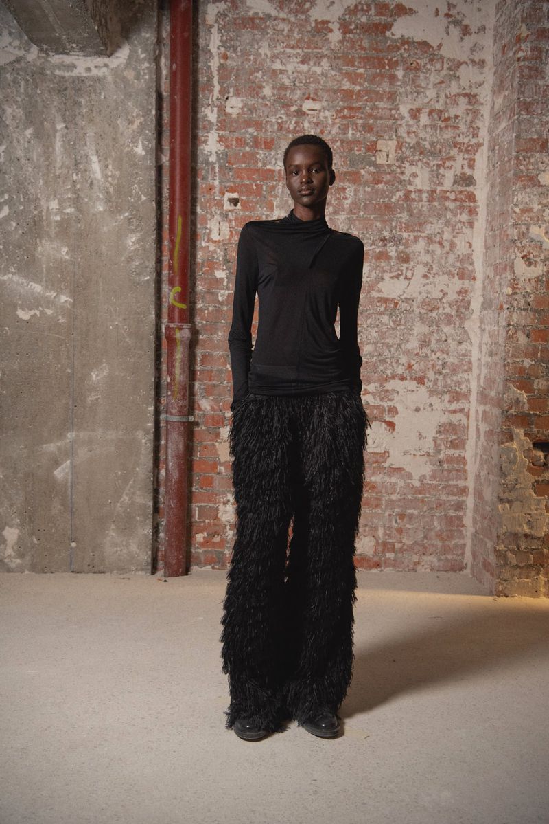An African-American fashion model in a long-sleeve top and feather pants stands in a studio with a brick wall backdrop.