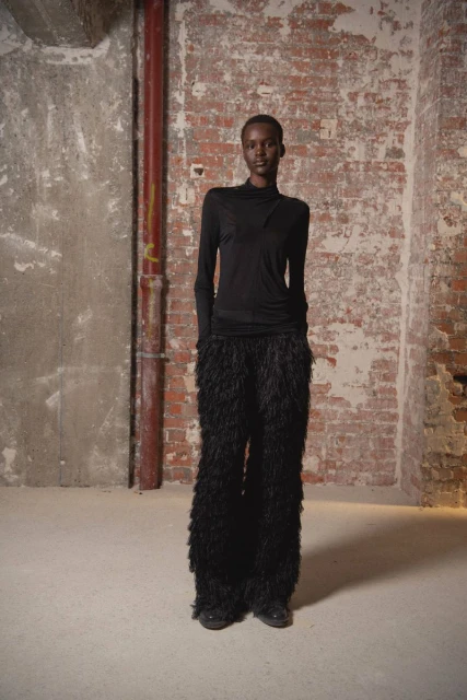 An African-American fashion model in a long-sleeve top and feather pants stands in a studio with a brick wall backdrop.