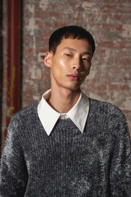 Asian man with short hair wearing a fuzzy grey sweater and white collared shirt, posing in front of a brick wall indoors.