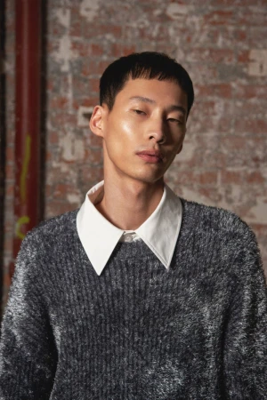 Asian man with short hair wearing a fuzzy grey sweater and white collared shirt, posing in front of a brick wall indoors.