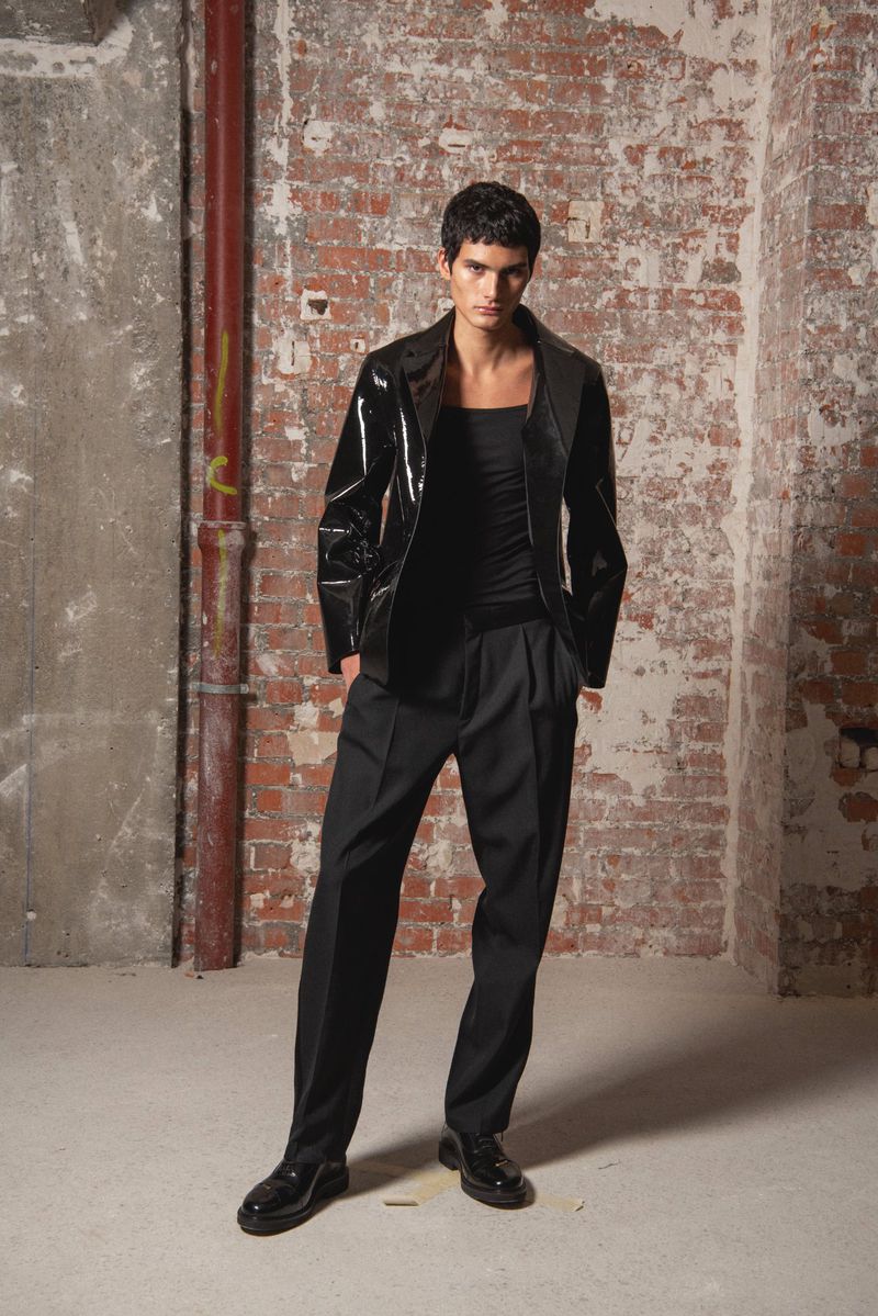 A slender man in a black patent leather jacket and pleated pants poses against a distressed brick wall in a full shot.