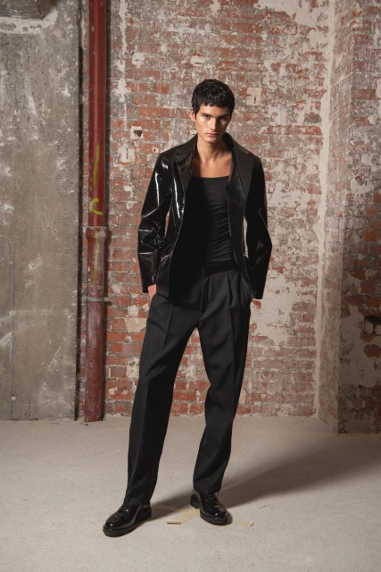 A slender man in a black patent leather jacket and pleated pants poses against a distressed brick wall in a full shot.