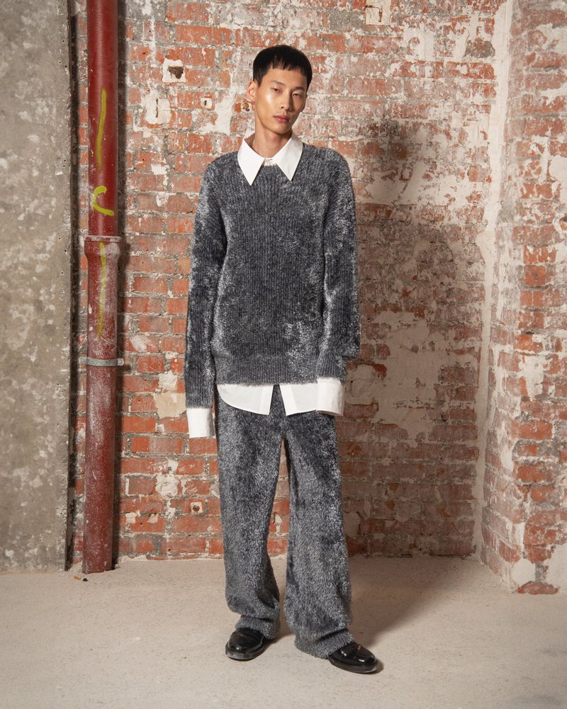 An Asian adult male model poses indoors, wearing a grey fluffy sweater and matching pants, along with black shoes. Red brick wall background.