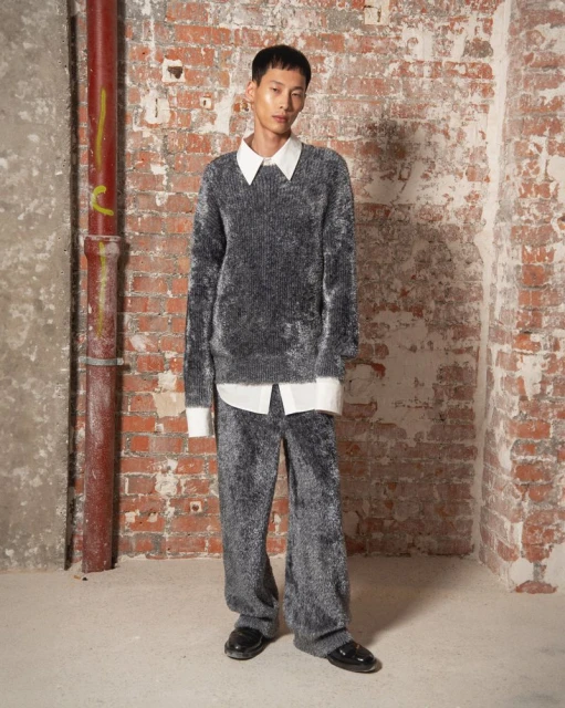 An Asian adult male model poses indoors, wearing a grey fluffy sweater and matching pants, along with black shoes. Red brick wall background.