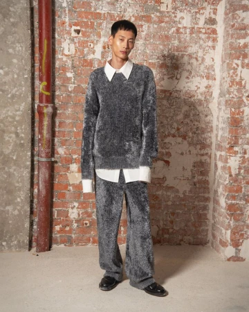 An Asian adult male model poses indoors, wearing a grey fluffy sweater and matching pants, along with black shoes. Red brick wall background.