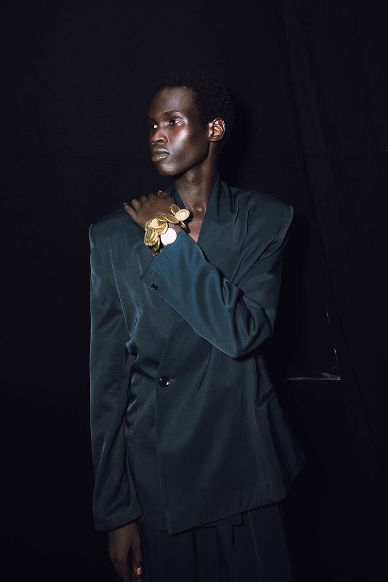 Portrait of an African American adult male wearing a teal suit with a large gold bracelet against a dark background in a studio.