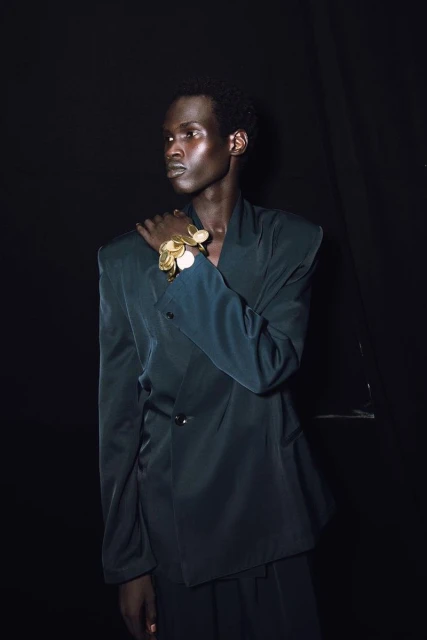 Portrait of an African American adult male wearing a teal suit with a large gold bracelet against a dark background in a studio.
