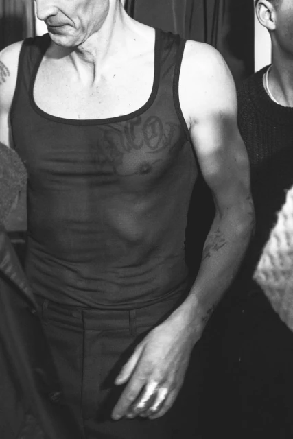 Monochrome portrait of shirtless adult male with tattoos, wearing a tank top, and standing indoors near another person.