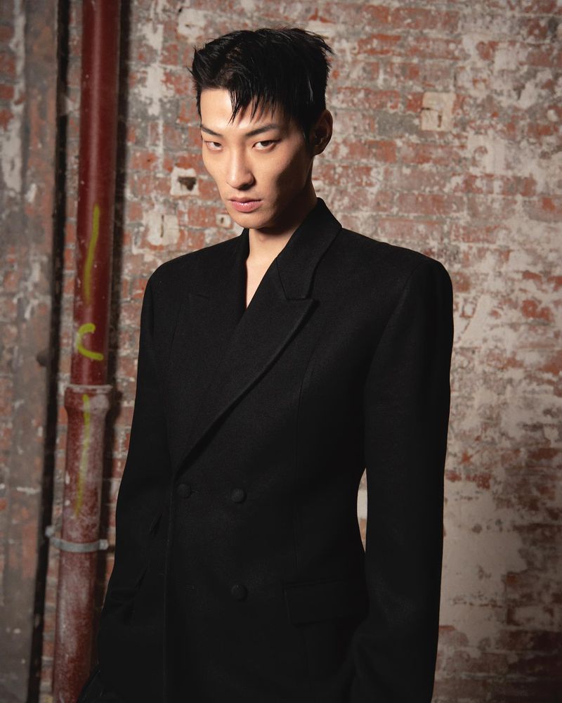 Close-up studio shot of a young Asian male model in a black double-breasted blazer against a distressed brick wall background.