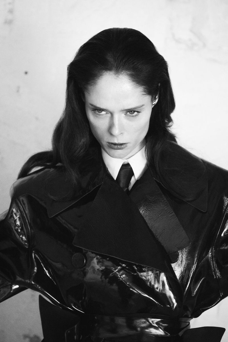 Monochrome portrait of adult female with dark hair in dark patent-leather-like double-breasted coat, looking down with a serious facial expression.