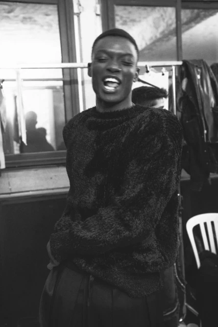A Black male model smiles widely in a backstage dressing room, wearing a textured black sweater and black pants, photographed in black and white.