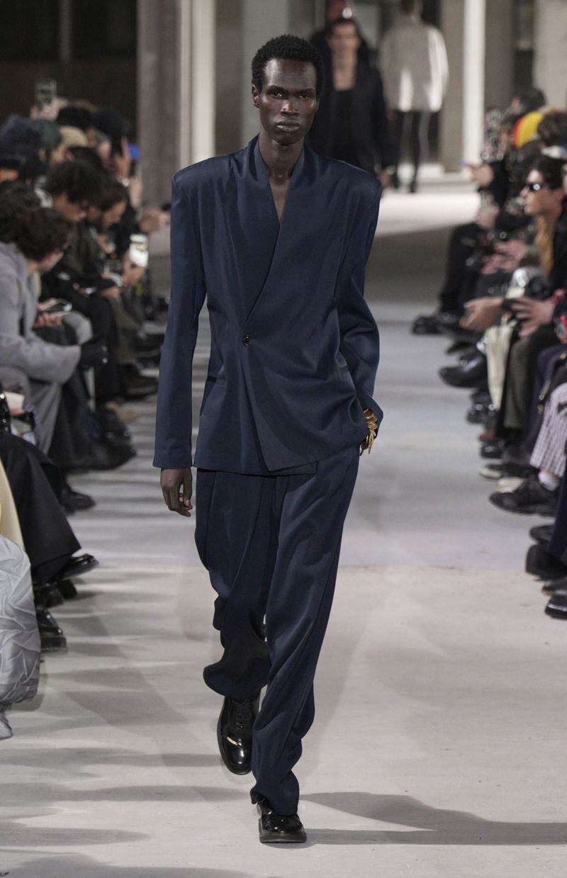 Runway fashion show features a male model of African descent wearing a tailored navy-blue suit and glossy black dress shoes, walking confidently.