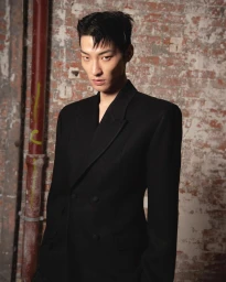 Model Xu Meen wearing a black tailored double-breasted coat against a weathered exposed brick wall backstage.