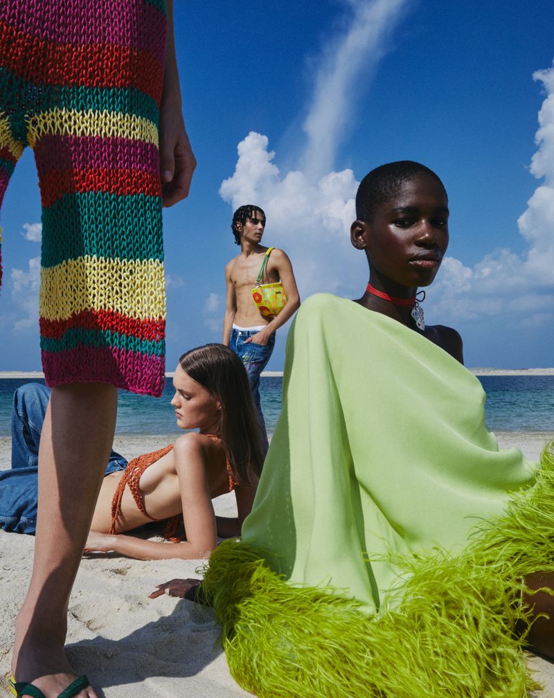 Fashion editorial shot on a beach featuring three models wearing colorful garments in a soft, bright light, complemented by a blue sky.
