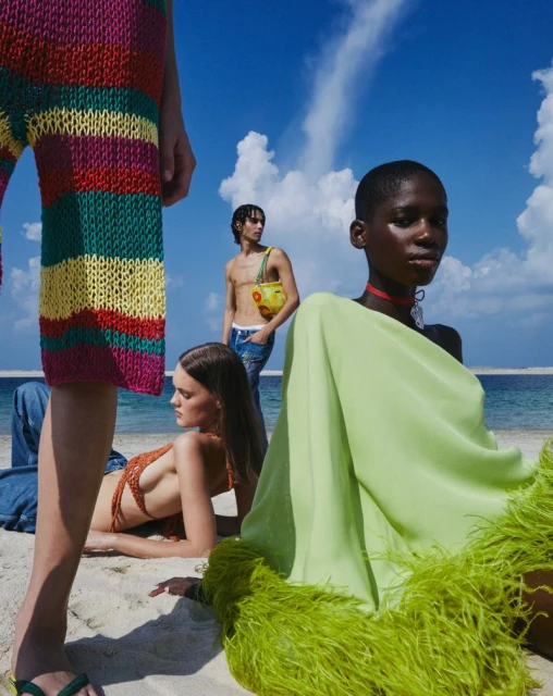 Fashion editorial shot on a beach featuring three models wearing colorful garments in a soft, bright light, complemented by a blue sky.