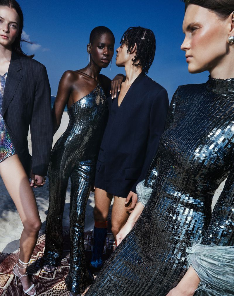 Four fashion models pose outdoors on a patterned rug. Three women wear sparkling outfits, while one man wears a dark blazer and shorts.