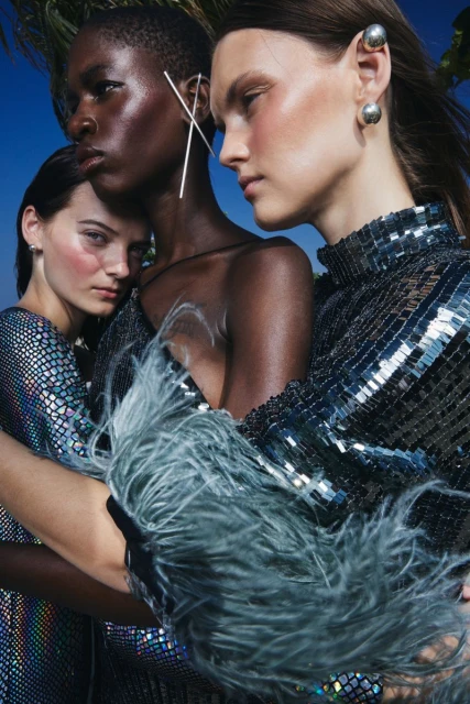 Three women in iridescent and sequined dresses pose closely against a blue sky, showcasing fashion earrings and iridescent makeup.
