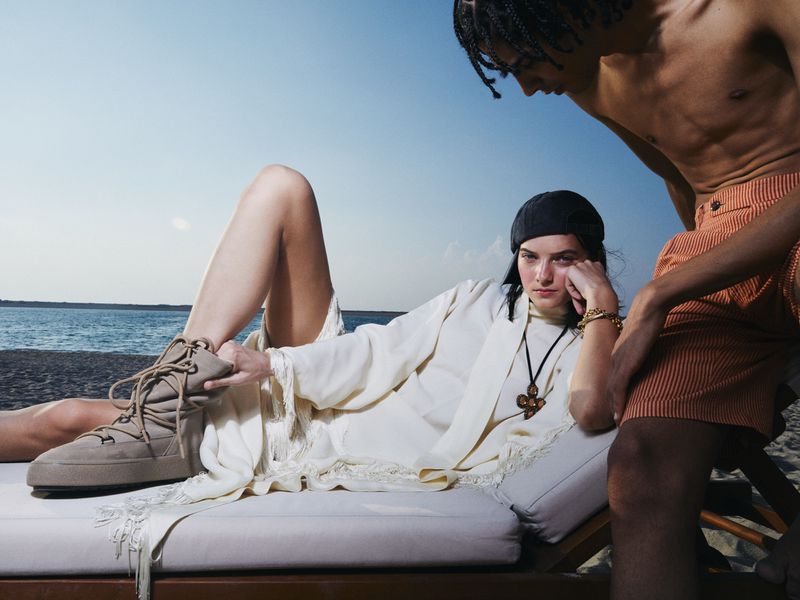 Two models on a beach lounge. One reclines wearing beige boots and a cream robe, while the other stands alongside in orange shorts.