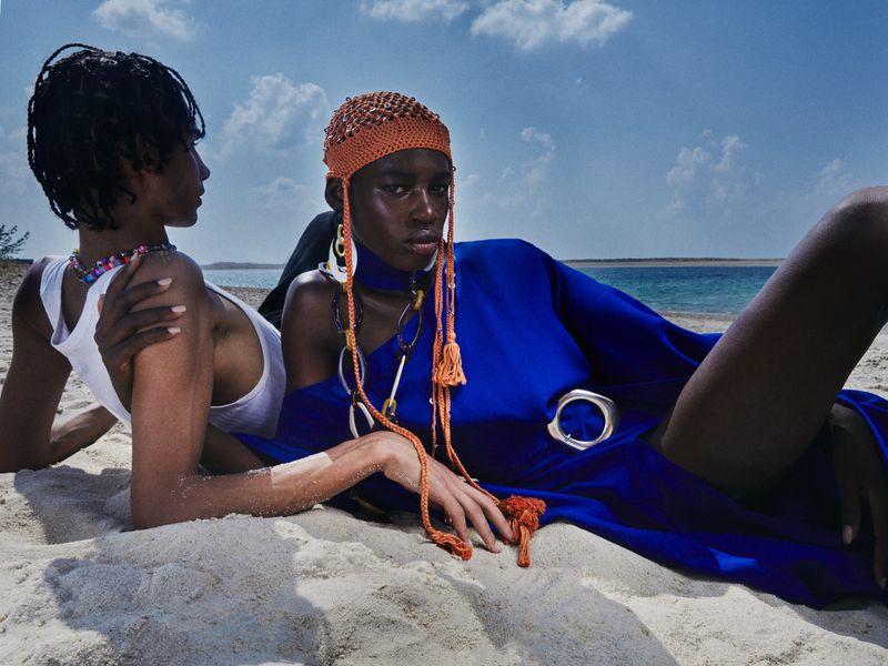 Two models resting on a sandy beach, one wearing a vibrant royal blue one-shoulder dress and an orange beaded knit headpiece.
