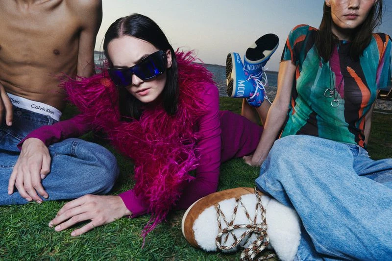 Three people lounge on the grass; a woman in purple with a feathered boa, a man with a bare torso, and a woman in a patterned shirt.