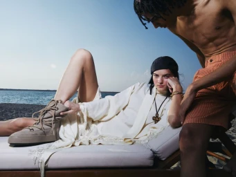 Woman reclining in white tunic adjusting beige lace-up boot on lounger by water while shirtless man stands nearby