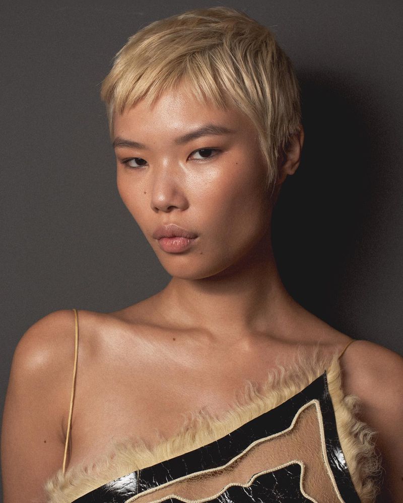 Close-up studio portrait of a young Asian woman with short blonde hair wearing a fur-trimmed dress with shoulder straps against a dark background.